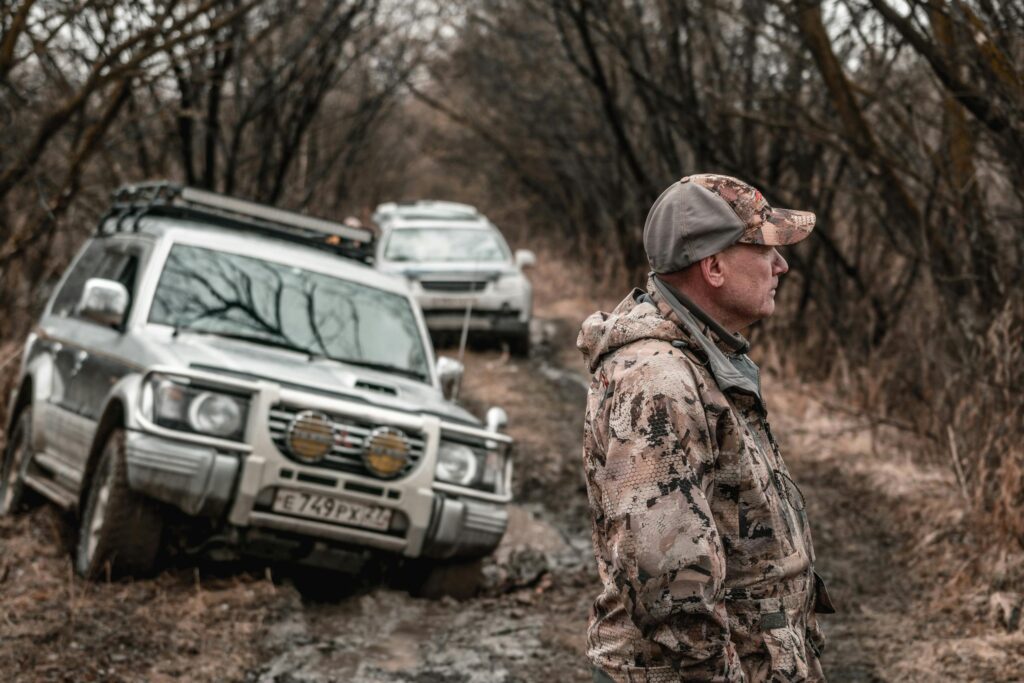 A man in camouflage stands by stuck off-road vehicles in a muddy forest trail.