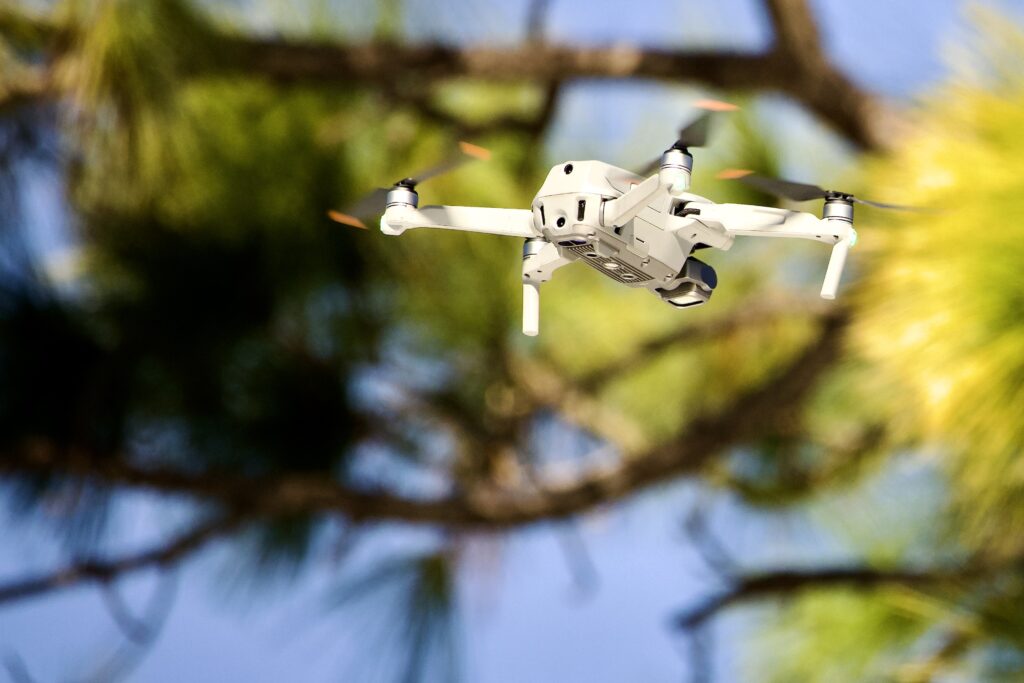 Professional photography drone hovering in a blue Florida sky, framed by pine trees, representing aerial industrial and real estate operations.